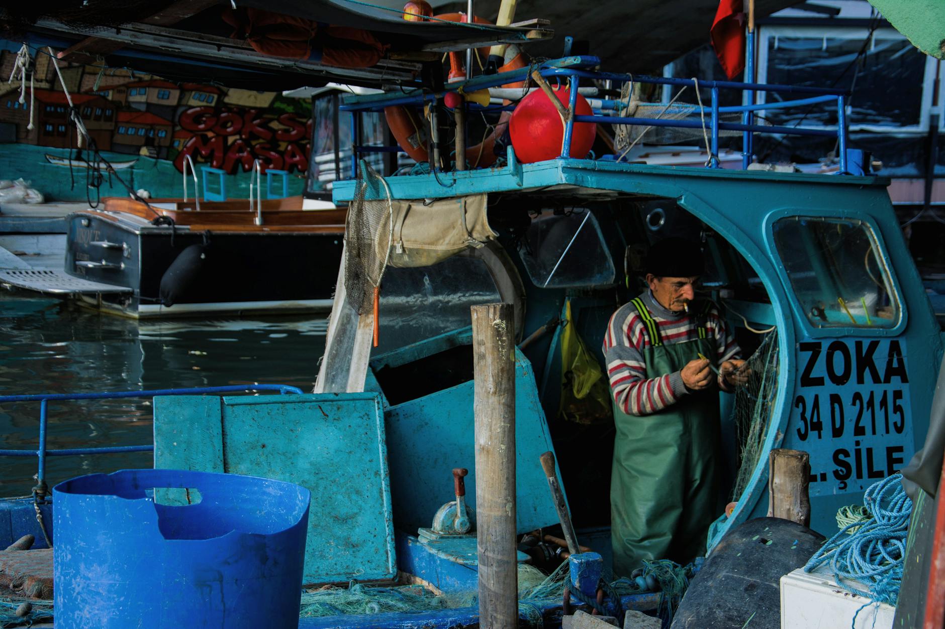 man in green overall smoking while fixing fishing net in old blue boat